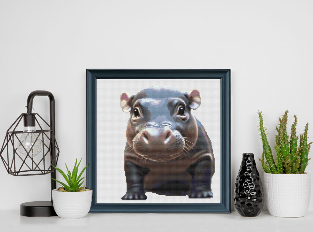 Baby pygmy hippo cross stitch pattern inspired by Moo Deng, stitched on white Aida cloth in a black frame