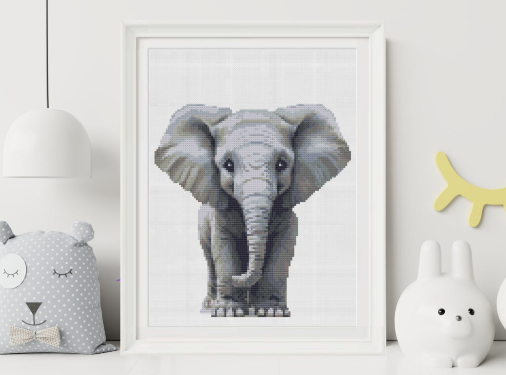 Cute elephant cross stitch on white Aida cloth in white fram sitting on a shelf with baby toys.